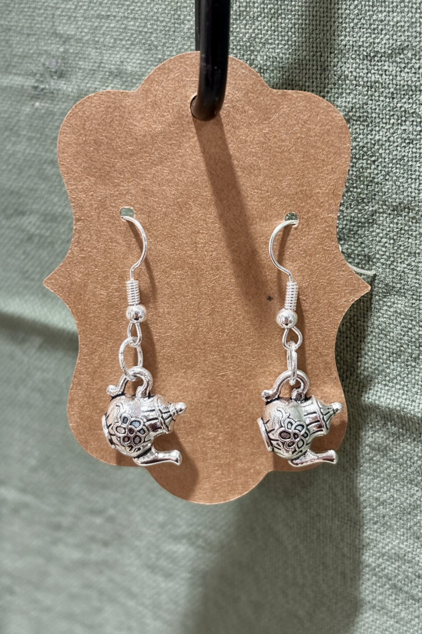 Silver teapot-shaped earrings on a brown earring card with a green fabric background